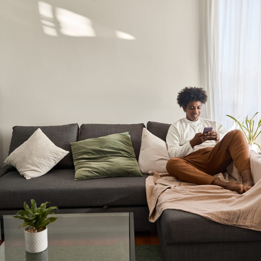 Happy relaxed gen z African American teen sitting on couch at home holding cellphone, using mobile apps on cell phone in modern living room. Social media influence concept. Authentic shot.